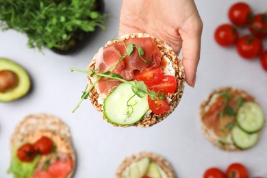 Woman holding crunchy buckwheat cakes with prosciutto, pieces of tomato and cucumber slice over white table, closeup