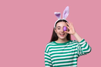 Photo of Happy woman in bunny ears headband holding painted Easter egg on pink background. Space for text.