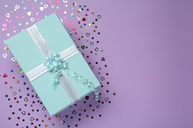 Light blue gift box and sequins on lilac background, flat lay. Space for text