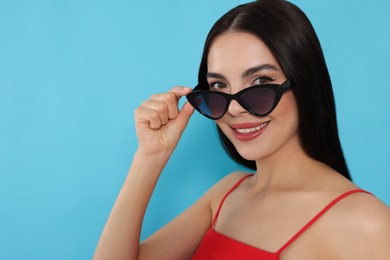 Attractive happy woman touching fashionable sunglasses against light blue background