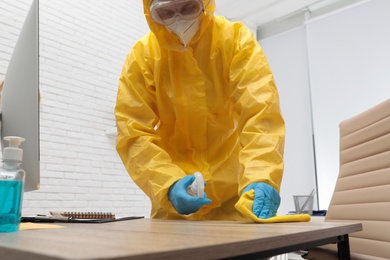 Janitor in protective suit disinfecting office furniture to prevent spreading of COVID-19