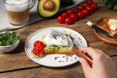 Photo of Woman eating delicious toast with poached egg and avocado at wooden table, closeup