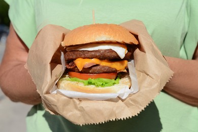 Photo of Woman holding delicious burger in paper wrap on blurred background, closeup