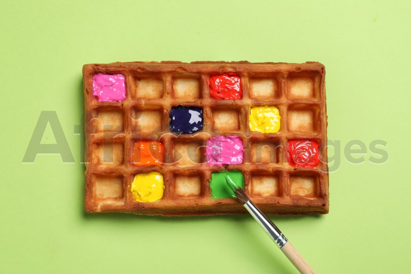 Waffle with different paints and brush on light green background, top view