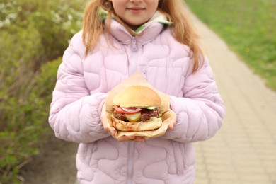 Little girl holding fresh delicious burger outdoors, closeup. Street food