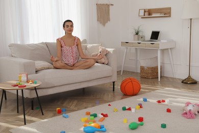 Photo of Calm young mother meditating on sofa in messy living room
