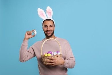 Photo of Happy African American man in bunny ears headband holding wicker basket with Easter eggs on light blue background. Space for text