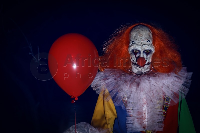Photo of Terrifying clown with red air balloon outdoors at night. Halloween party costume