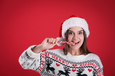 Photo of Emotional woman in Santa hat and Christmas sweater eating candy cane on red background