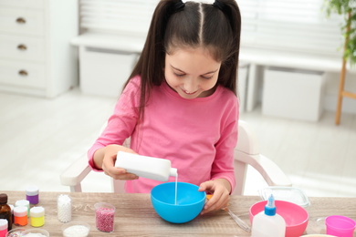 Cute little girl pouring glue into bowl at table in room. DIY slime toy
