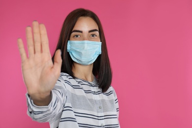 Young woman in protective mask showing stop gesture on pink background, space for text. Prevent spreading of coronavirus