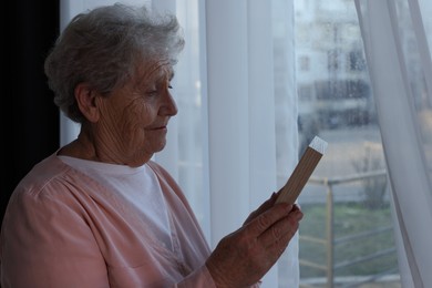 Elderly woman with photo frame near window indoors. Loneliness concept