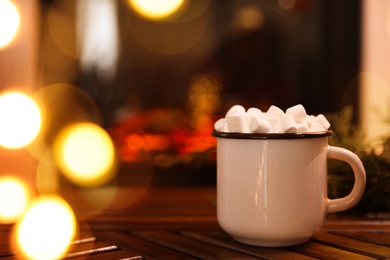Cup of hot drink with marshmallows on wooden table near fireplace at home. Space for text