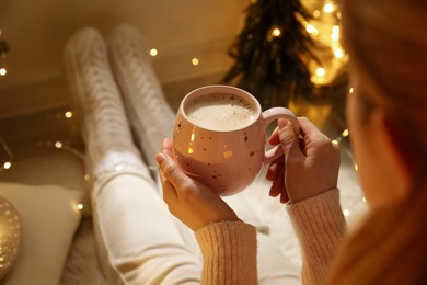 Woman holding cup with hot drink near festive lights at home, closeup