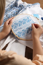 Woman embroidering white shirt with blue thread at wooden table, closeup. Ukrainian national clothes