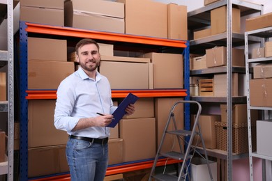 Young businessman with clipboard near rack of cardboard boxes at warehouse