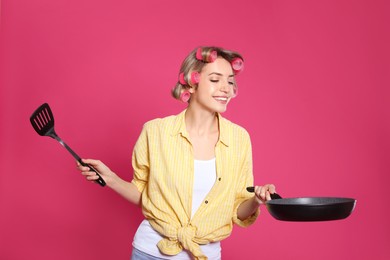 Young housewife with frying pan and spatula on pink background