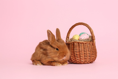 Adorable fluffy bunny near wicker basket with Easter eggs on pink background
