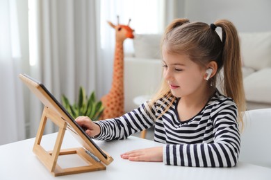 Photo of Adorable little girl doing homework with tablet and earphones at table indoors