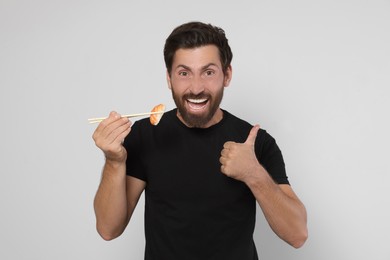 Emotional man holding tasty sushi with chopsticks and showing thumbs up on light grey background