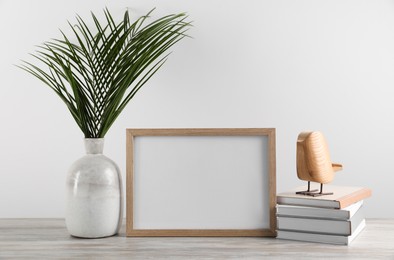 Blank photo frame, books, decorative bird and tropical leaves on wooden table