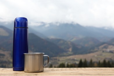 Blue thermos and metal mug on wooden table against mountain landscape. Space for text