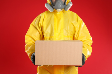 Man wearing chemical protective suit with cardboard box on red background, closeup. Prevention of virus spread