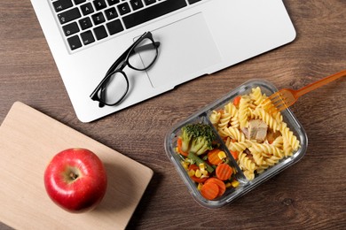 Photo of Container of tasty food, laptop, apple, book and glasses on wooden table, flat lay. Business lunch