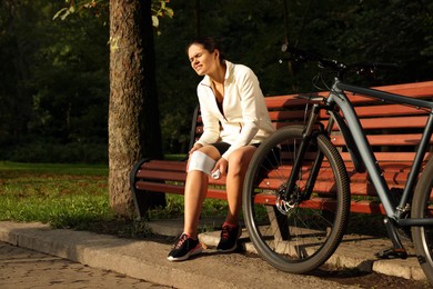 Young woman applying bandage onto her knee on wooden bench outdoors