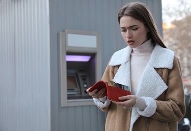 Photo of Unhappy young woman with wallet near cash machine outdoors