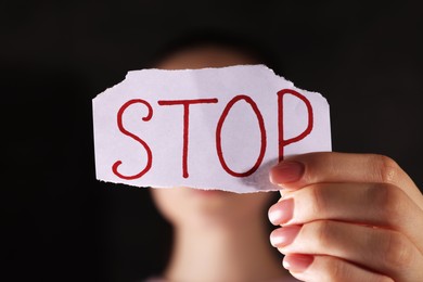 Photo of Woman holding piece of paper with word Stop on dark background, closeup. Domestic violence concept