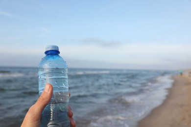 Photo of Woman holding plastic bottle with water near sea, closeup. Space for text