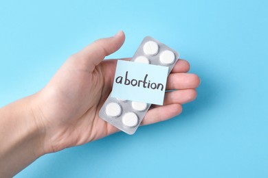 Woman holding paper note with word Abortion and pills on light blue background, top view