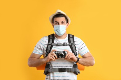 Male tourist in protective mask with travel backpack and camera on yellow background