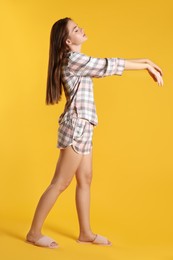 Young woman wearing pajamas and slippers in sleepwalking state on yellow background