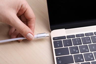Woman plugging USB cable with type C connector into laptop port on light wooden table, closeup