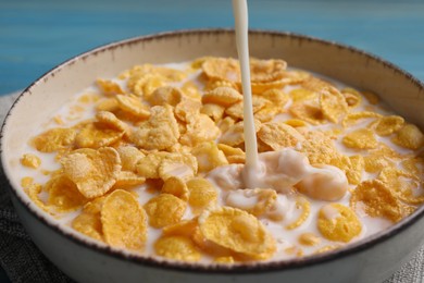 Pouring milk into bowl of tasty corn flakes at light blue table, closeup