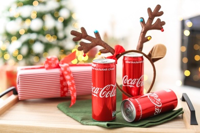 MYKOLAIV, UKRAINE - January 01, 2021: Cans of Coca-Cola and gift on tray against blurred Christmas lights