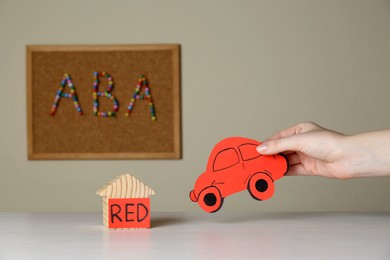 Photo of Woman holding red car near wooden house and cork board with abbreviation ABA (Applied behavior analysis) at white table, closeup