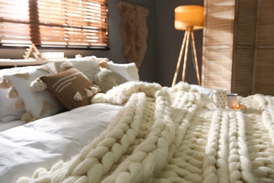 Bed with cozy knitted blanket and cushions near window in room. Interior design
