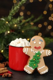 Photo of Gingerbread man and red cup with marshmallows on black table against blurred festive lights. Making homemade Christmas cookies