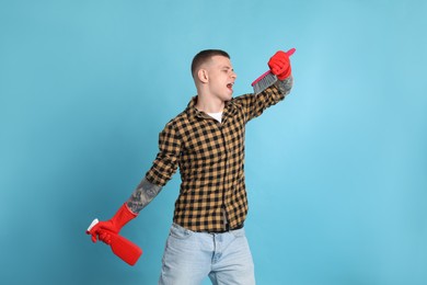 Handsome young man with brush and bottle of detergent singing on light blue background