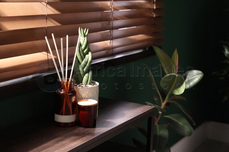 Reed freshener, candle and houseplant on table near window indoors