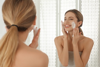 Young woman applying cleansing foam onto her face near mirror in bathroom