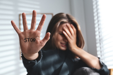 Crying young woman showing palm with word STOP indoors, focus on hand. Domestic violence concept