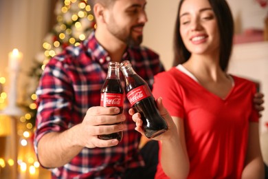 Photo of MYKOLAIV, UKRAINE - JANUARY 27, 2021: Young couple holding bottles of Coca-Cola in room decorated for Christmas, focus on hands