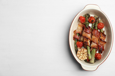 Oven baked asparagus wrapped with bacon in ceramic dish on white wooden table, top view. Space for text
