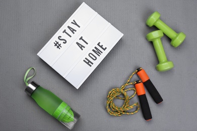 Sport equipment and lightbox with hashtag STAY AT HOME on grey yoga mat, flat lay. Message to promote self-isolation during COVID‑19 pandemic