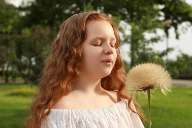 Cute girl with beautiful red hair blowing large dandelion in park. Allergy free concept
