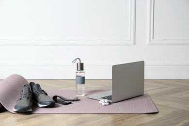 Exercise mat, laptop, bottle of water, wireless earphones, fitness elastic band and shoes on wooden floor indoors